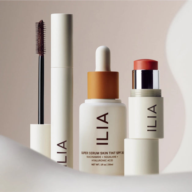 ILIA beauty products arranged on a neutral surface, including mascara, face milk, foundation, and a multi-stick with flowers in the background.