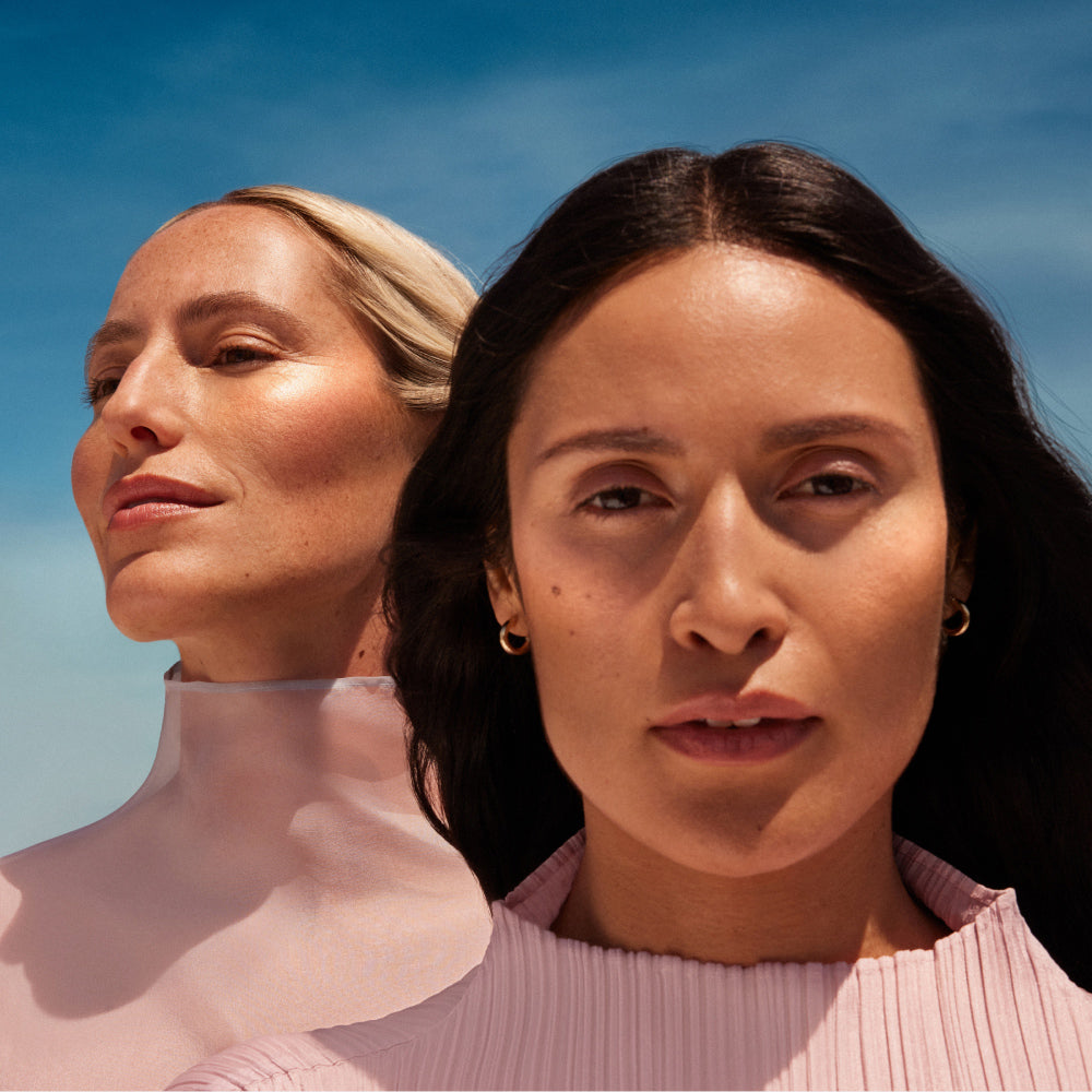Two models with bronzy and glowy skin on a sunny day in front of a sky of light clouds.