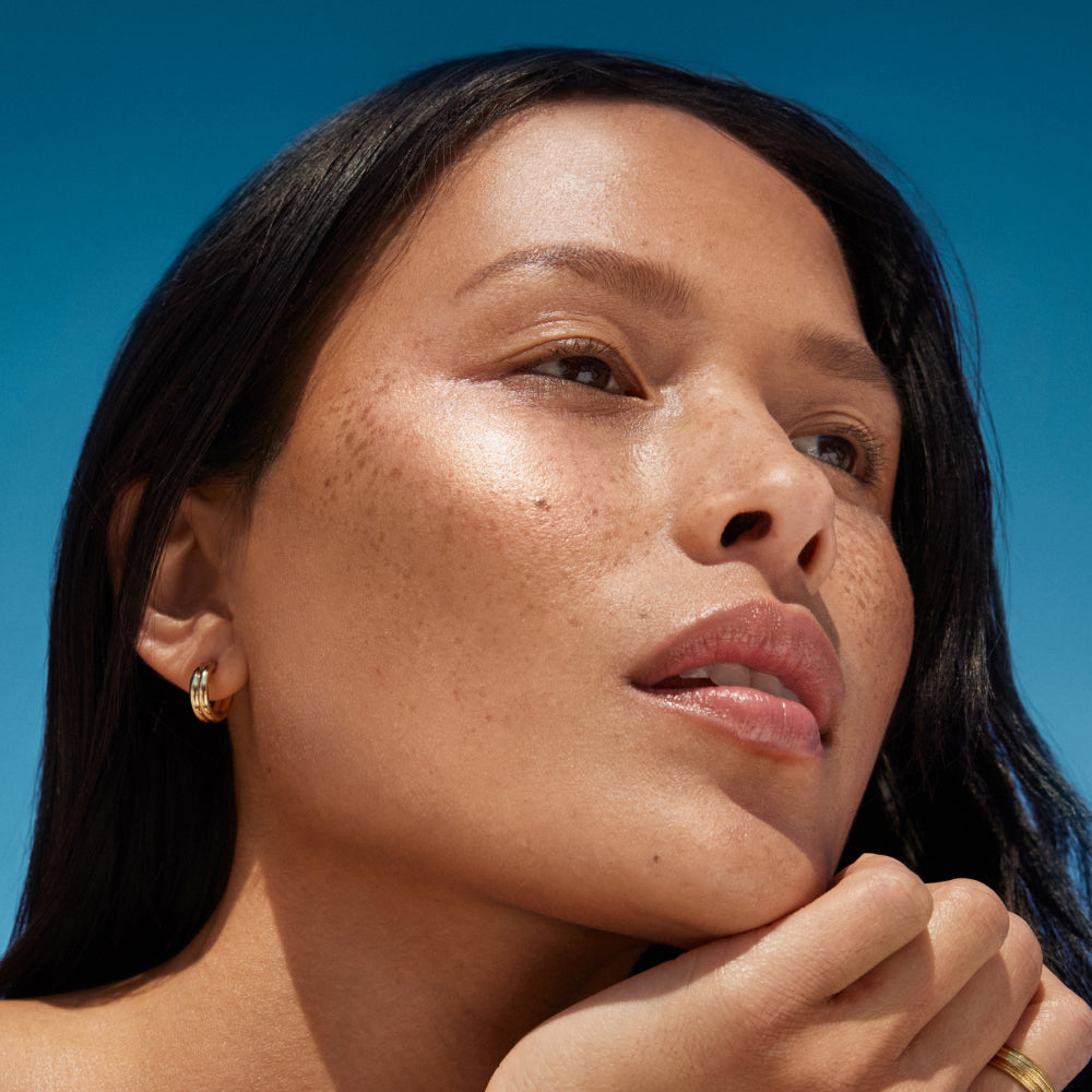 A freckled model with bronzy, glowy skin in a candid shot with a blue sky background.