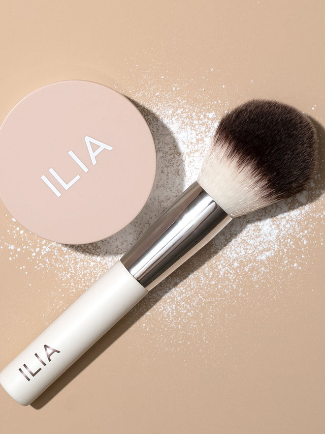An ILIA powder lying next to a domed, full-bodied brush on a tan background.