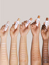 Hands holding ILIA Complexion Stick with swatches of all shades on skin against a neutral background