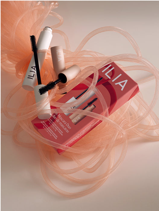 ILIA makeup products including two mascaras on a light background with sheer peach fabric.