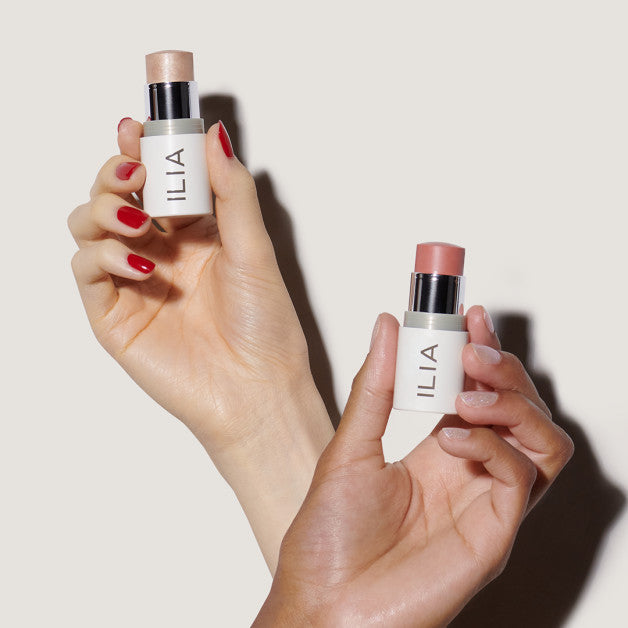 Two hands holding ILIA multi-use makeup sticks, one in a shimmery highlight and the other in a soft pink shade.