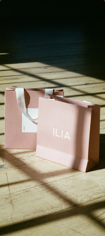 Two pink 'ILIA' shopping bags on a wooden floor with sunlight casting shadows.