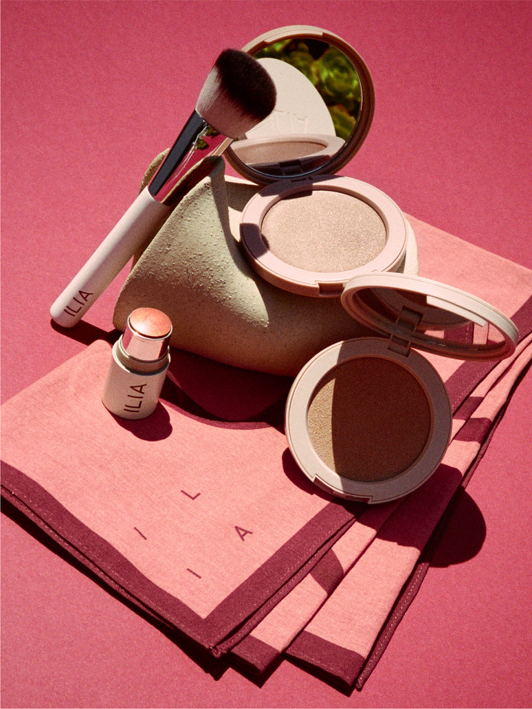 Makeup products including a compact, brush, and small lip + cheek stick on a pink surface with 'ILIA' branded on a scarf.