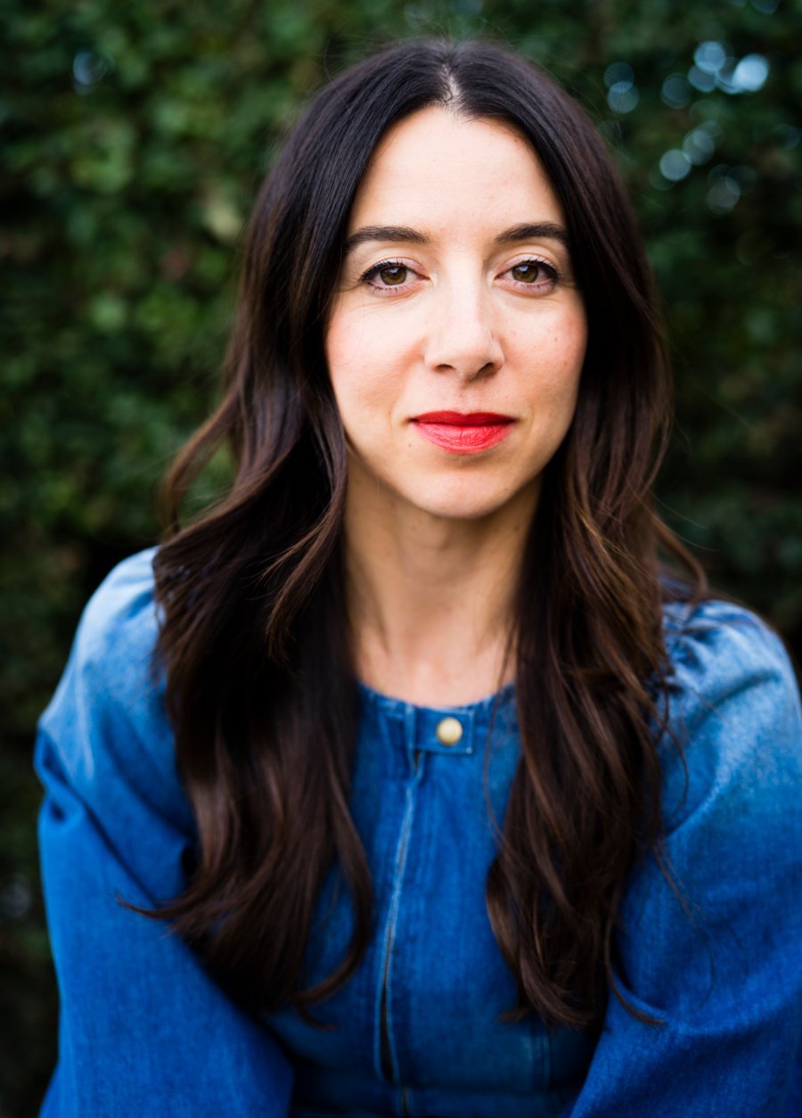 The founder of ILIA, Sasha, with a red lipstick and denim top looks directly into the camera with a confident expression against a blurred greenery background.